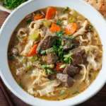 A steaming bowl of homemade beef noodle soup garnished with herbs and spices.
