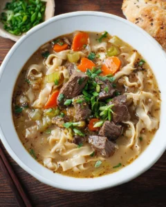 A steaming bowl of homemade beef noodle soup garnished with herbs and spices.